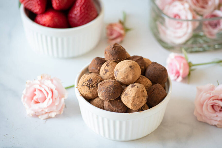 Adaptogen chocolate truffles dusted with cacao in a white bowl beside pink roses and strawberries.