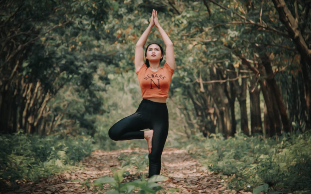 Woman in orange shirt and black pants in the forest doing tree pose during vata season.
