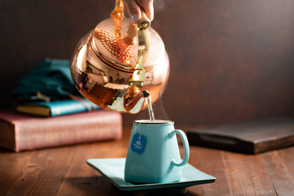 Pouring ayurveda tea from a copper pot into a teal teacup and saucer.