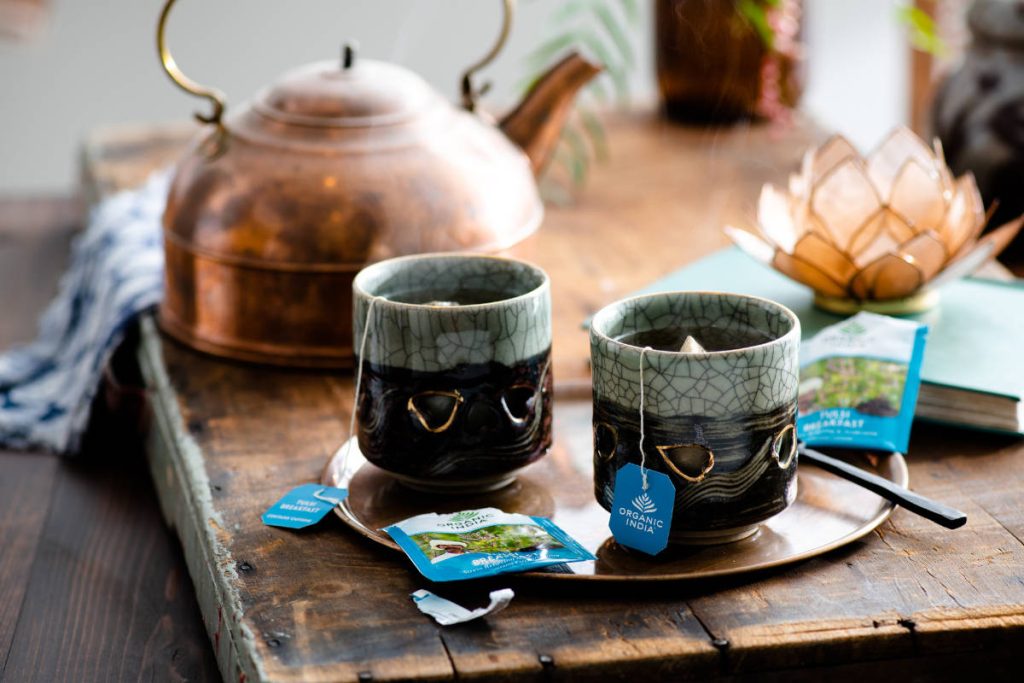 Tulsi breakfast tea in ceramic tea cops with copper teapot on wood table with lotus candle holder.