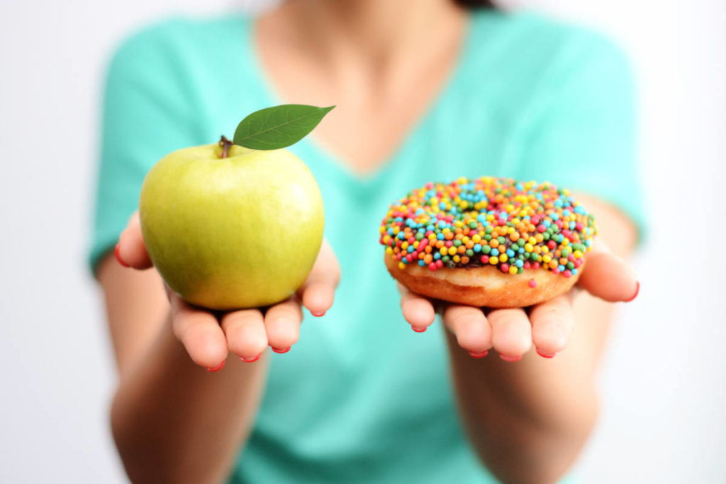 Woman holding a green apple in one hand and chocolate frosted donut with sprinkles in the other to choose between for blood sugar health.