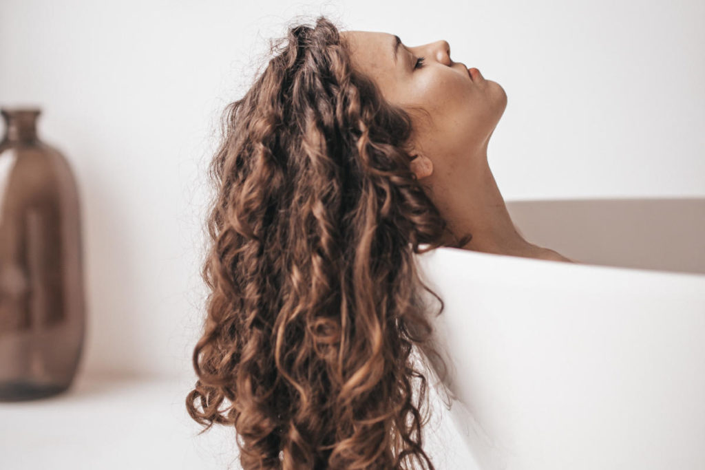 Women's brown curly hair hanging out of the edge of a white bathtub.