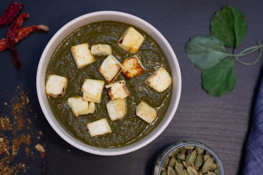 A bowl of saag paneer, or creamed spinach with cooked cheese, with a little bowl of cardamom seeds on the side. 