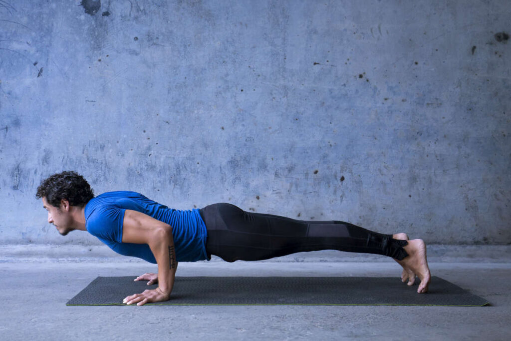 Man with blue shirt and black pants on black yoga mat doing chaturanga yoga pose for running.