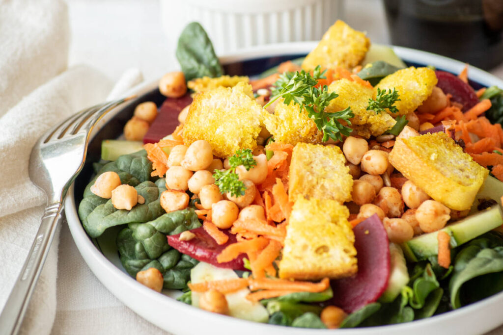 Homemade croutons with Turmeric over a mixed salad of chickpeas, spinach and beets in white bowl with fork.