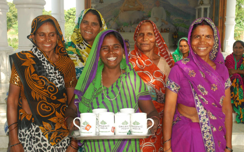 Indian women in saris mid-laugh holding a platter of Organic India tea cups.