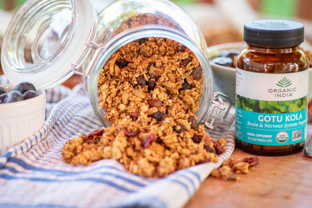 Gotu kola granola spilling out of a clear jar and a small bowl of blueberries.