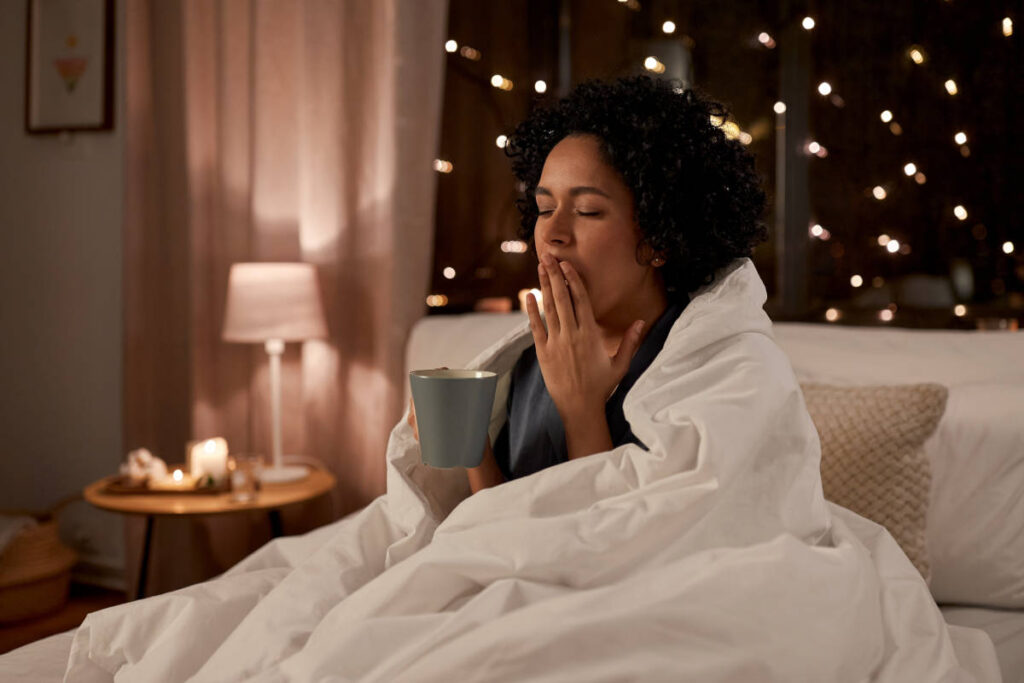 Woman in bed yawning while holding a cup of tea with herbs for sleep. 