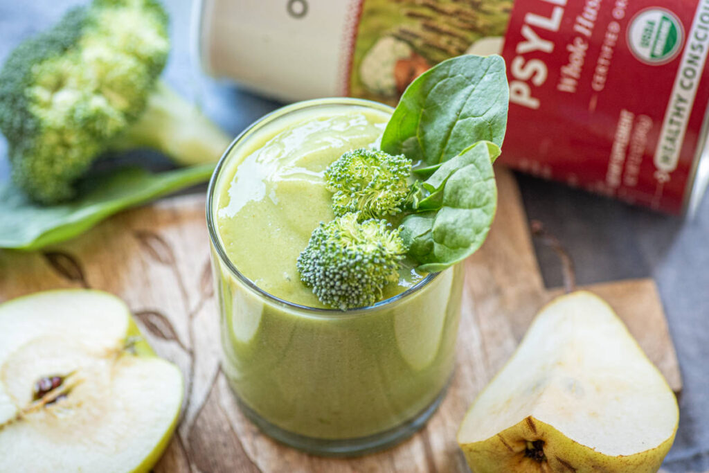 Green high fiber smoothie with broccoli and spinach garnish in a clear glass on a wood cutting board beside cut pears and apples.