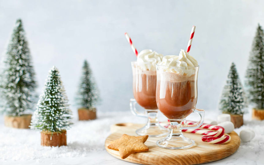 Chocolate holiday lattes on a cutting board with cookies, candy canes and figurine trees.