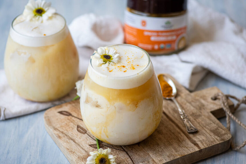 Iced golden milk spiced turmeric latte in glasses with foam, flower garnish and a scoop of vibrant turmeric mix.