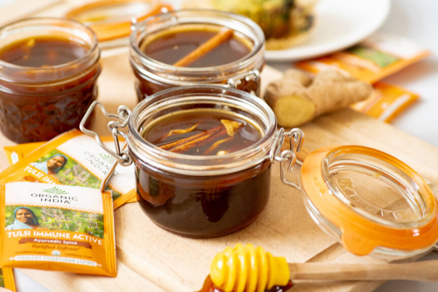 Jars of honey with herbs and spices on a cutting board with ginger and honey dipper.
