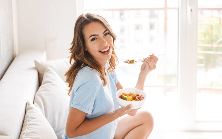 Woman sitting on a white couch smiling and eating a bowl of fruit to support adaptive and innate immunity.
