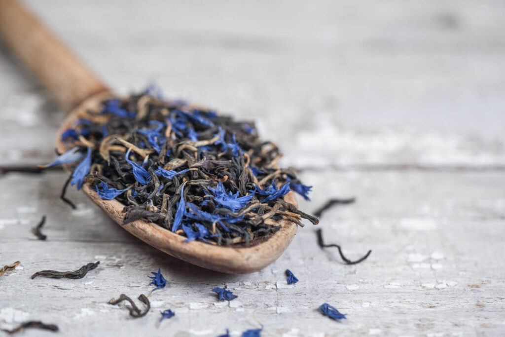 Loose leaf black tea and dried blue flowers on a wooden spoon on a wooden table.