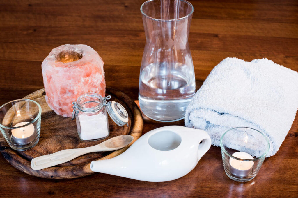 Neti pot for spring allergies on a wooden table with candle, hand towel, water, and Himalayan pink salt. 