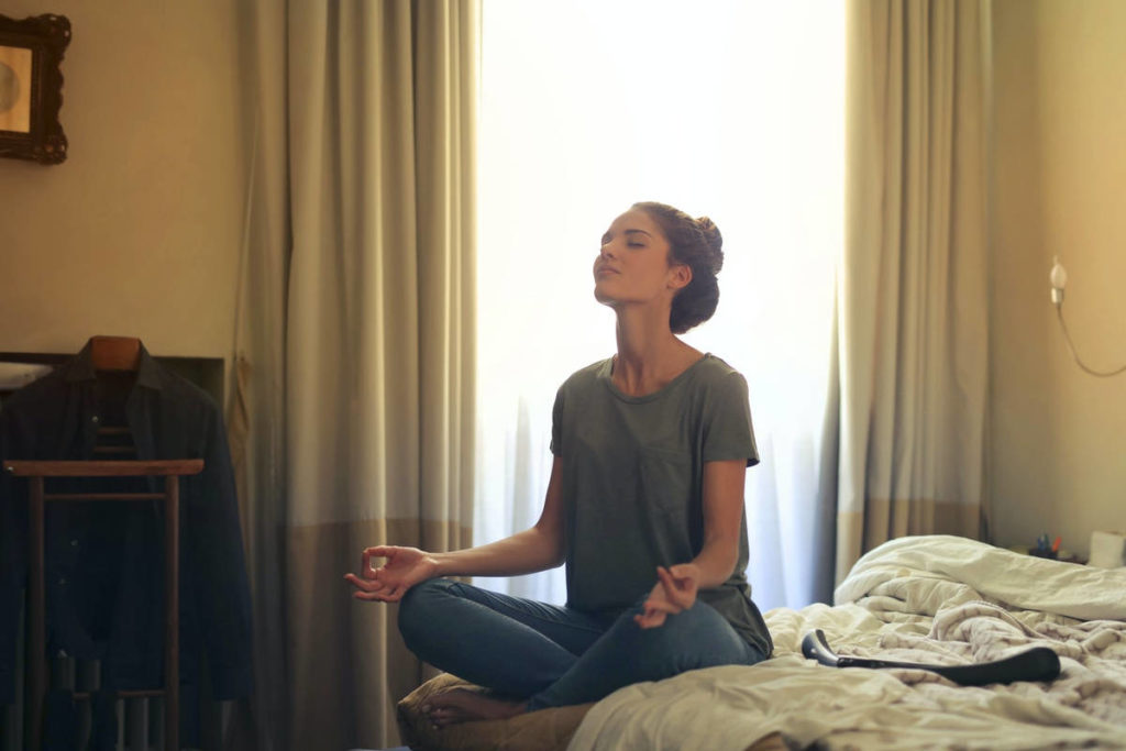 Women practicing pranayama meditation in bed with light coming through the window.