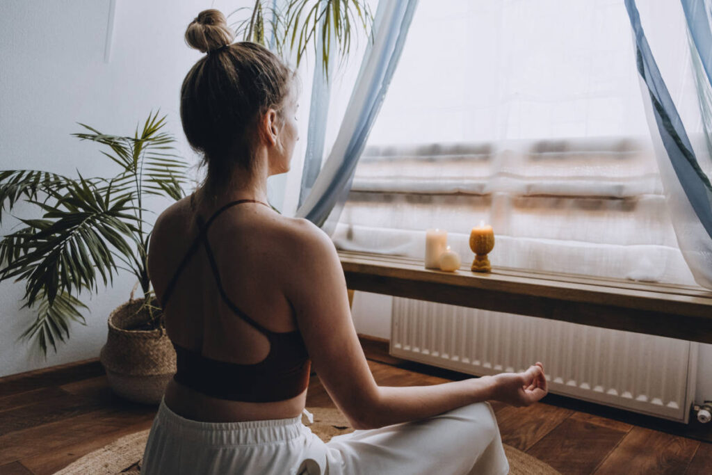 Woman practicing home pranayama meditation with candles and plants.