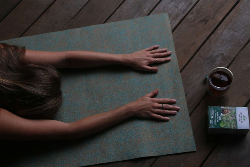 Woman comsuning rasayana herbs tulsi tea while doing child's pose on green mat on wood floor.