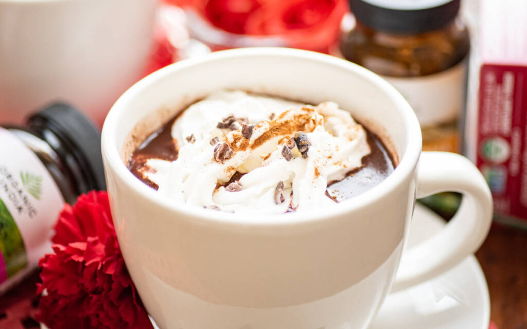 Cinnamon hot chocolate latte with rose and ashwagandha in a white mug topped with nondairy whip and cacao nibs.