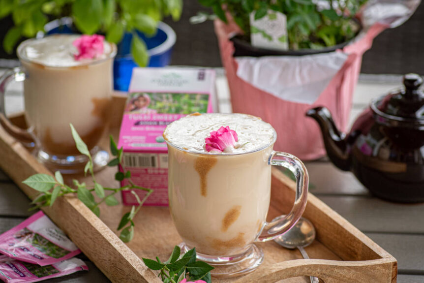 Creamy rose tea latte in a glass cup with caramel drizzle and rose garnish on wooden serving tray with tea kettle.