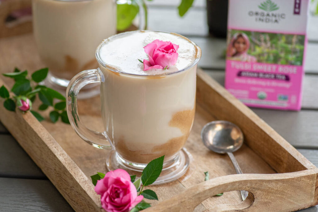 Creamy rose tea latte in a glass cup with caramel drizzle and rose garnish on wooden serving tray. 