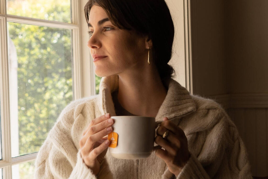 Woman practicing self nurturing by drinking tea in cozy sweater by the window.