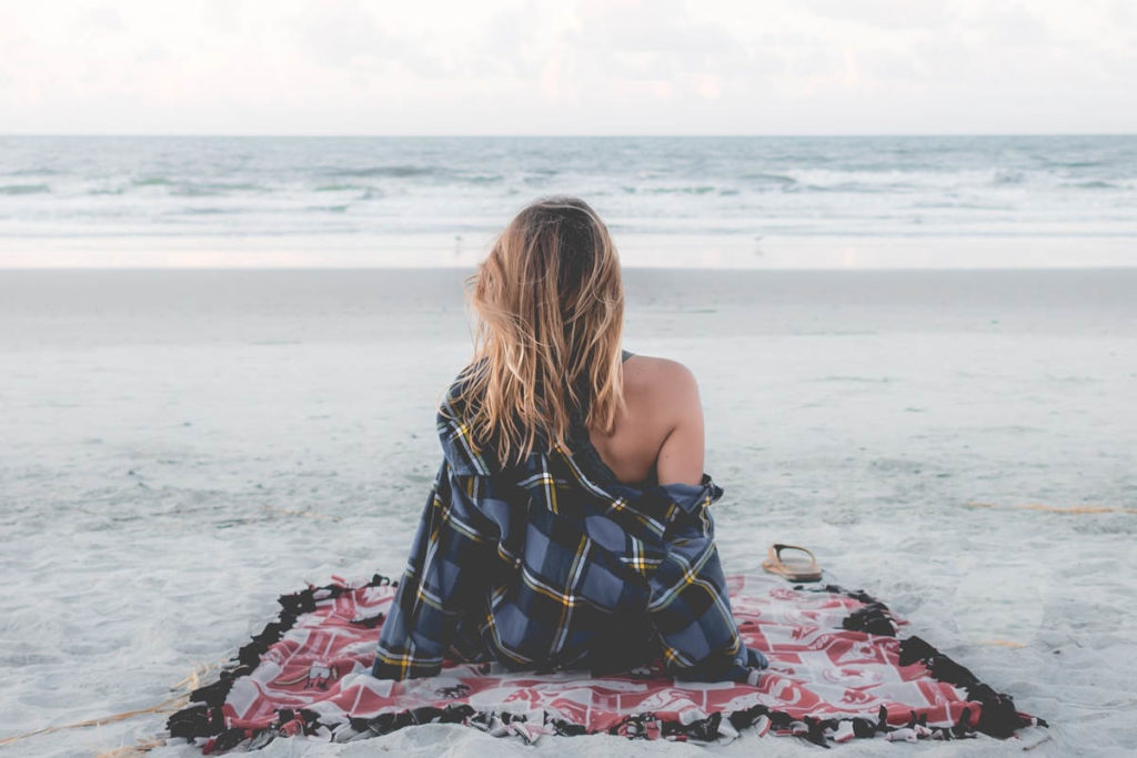 Woman with blond hair sitting on beach on checkered red blanket with plaid blue button down looking out to the ocean to set intention.