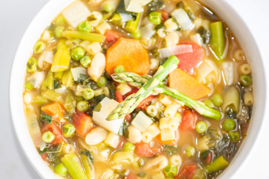 Spring minestrone soup with asparagus, ditalini, carrots, peas and kale in a white bowl. 