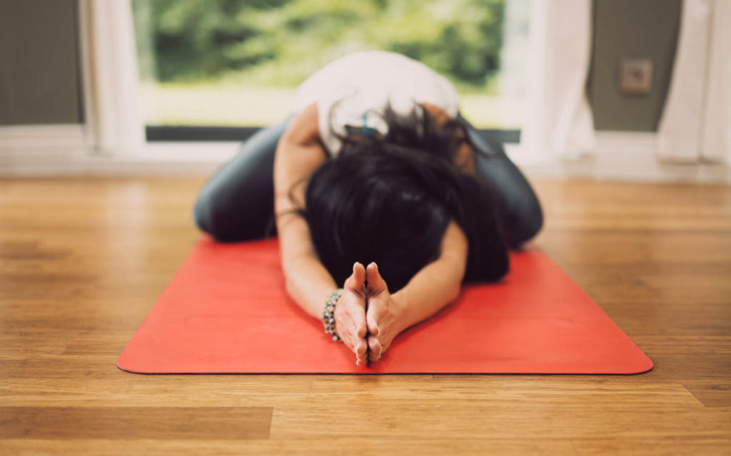 Woman doing childs pose with prayer hands on an orange yoga mat and wood floor. 