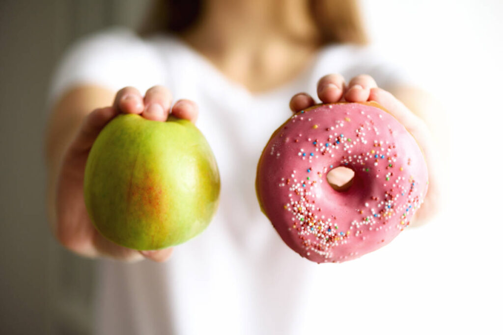 Sugar detox woman in white shirt holding a green apple in one hand and a strawberry frosted donut in the other.