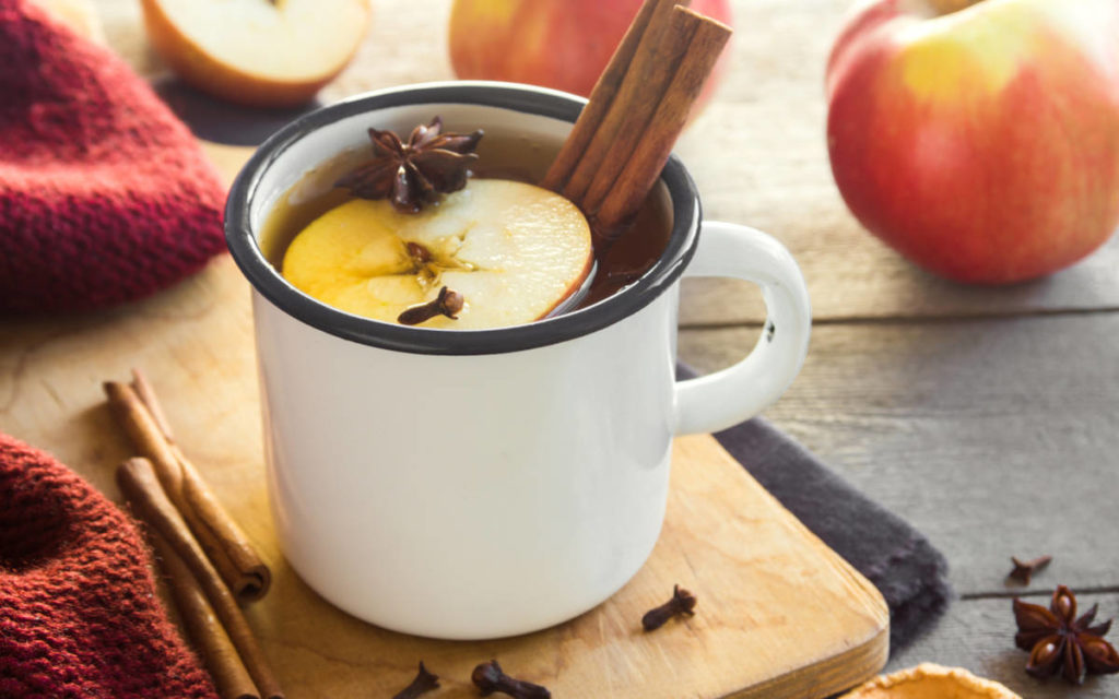Mug of hot apple cider with cinnamon sticks, clove, and fresh apple on a wooden table. 