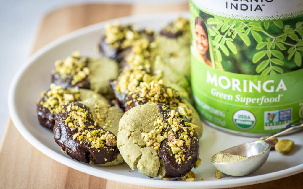 Moringa pistachio cookies on a white plate half dipped in chocolate with pistachio bits. 