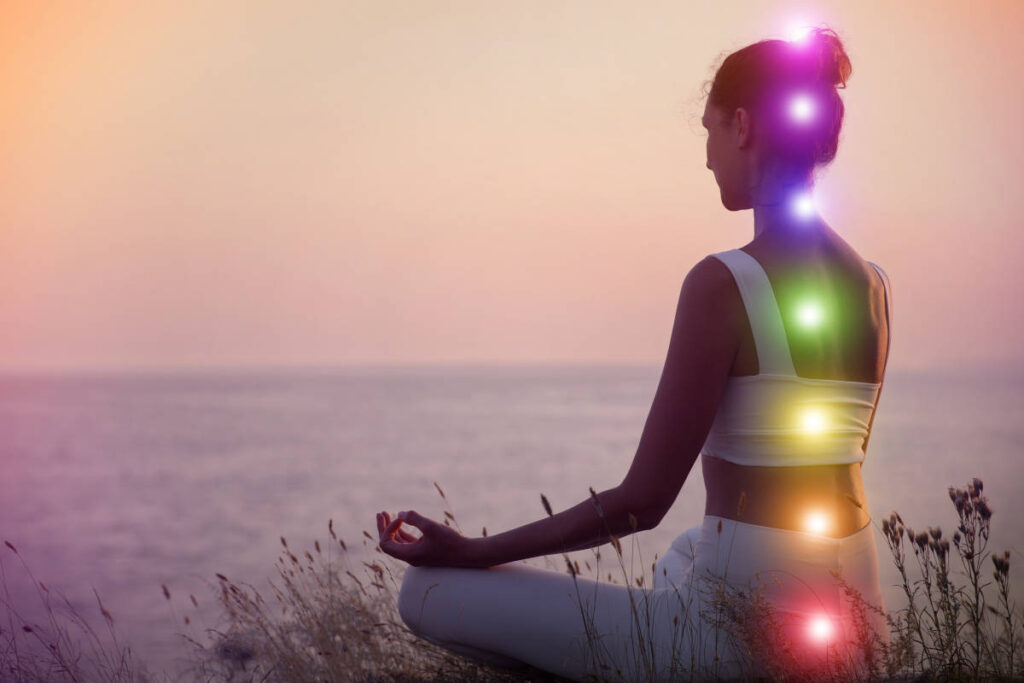 Woman wearing white sitting in the grass by the see with the 7 chakras illuminated in their correlating color.