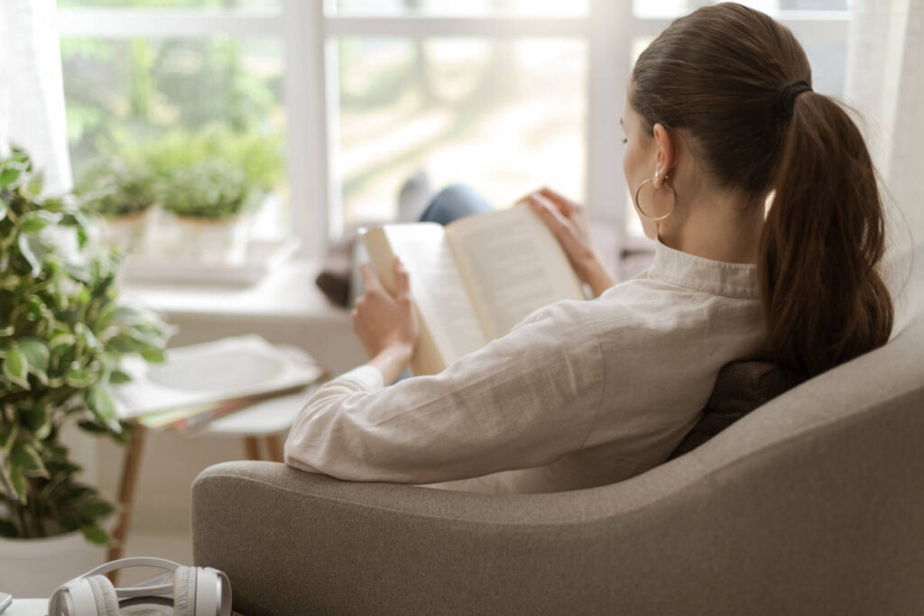 Woman with brown hair in ponytail and linen shirt reading a book with nervine herbs and houseplants nearby.