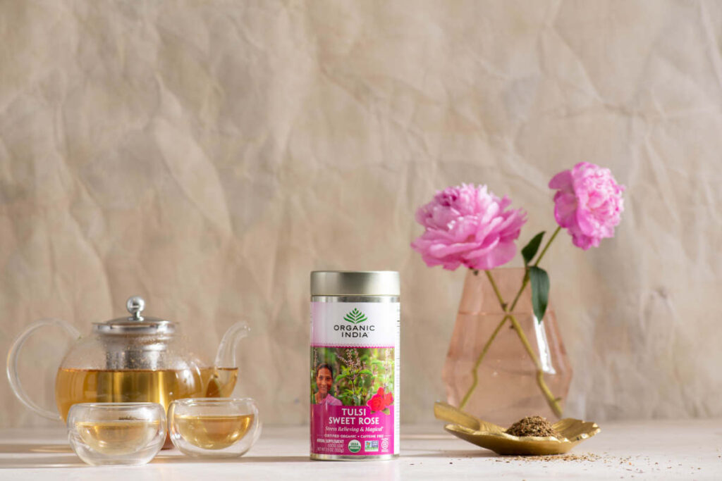 Tulsi Sweet Rose lloose leaf tea in canister and on a saucer with clear tea infuser teapot and cups.