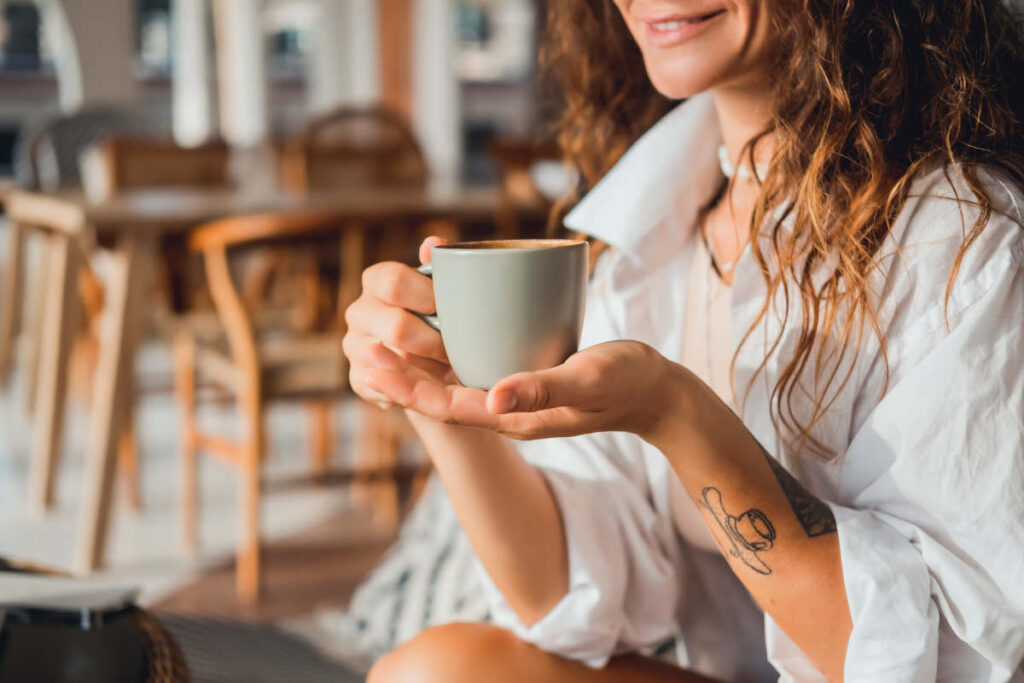 Woman with light brown curly hair and tattoos holding a grey ceramic tea mug.