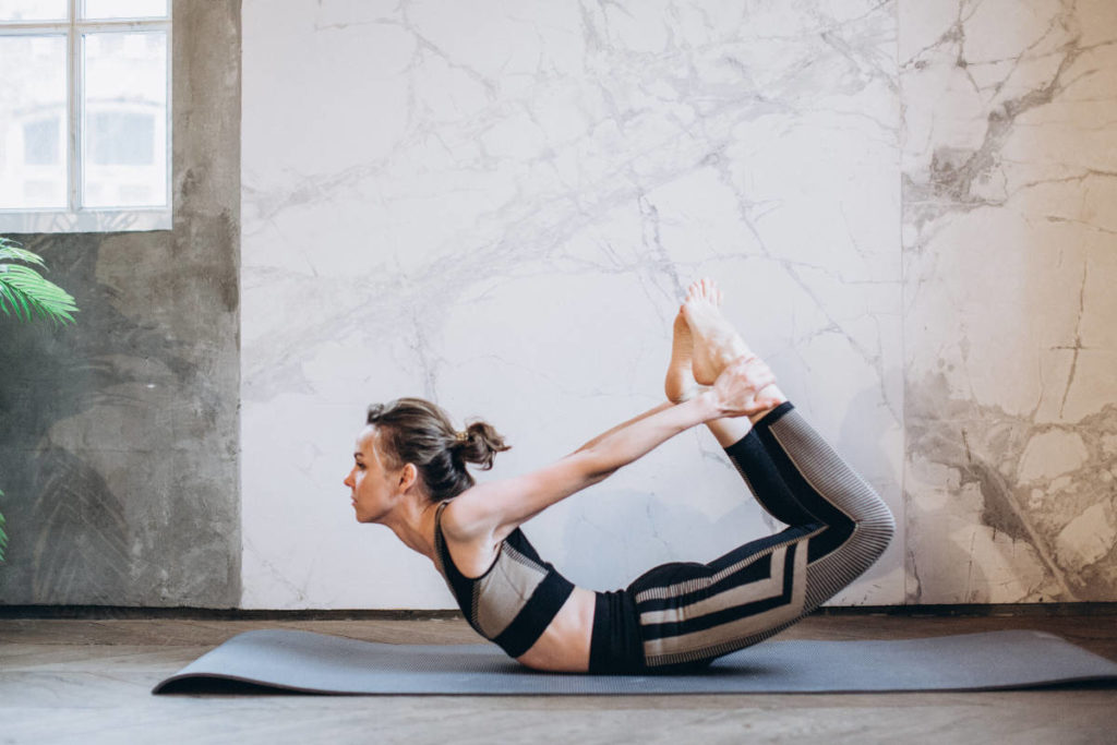 Woman in black and tan yoga outfit and messy bun doing bow pose on grey yoga mat.