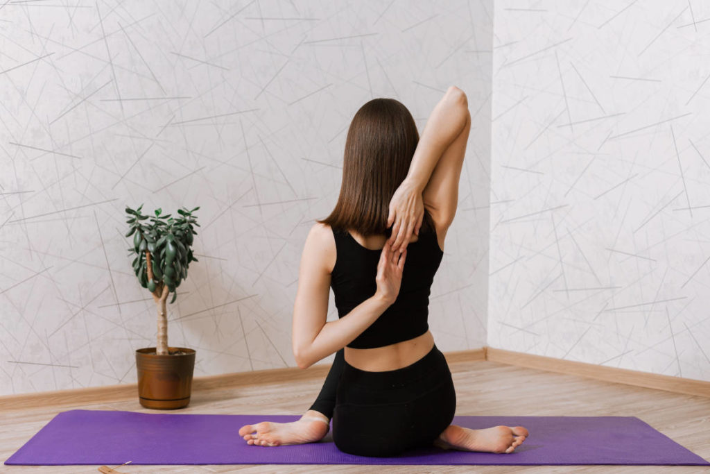 Woman in all black on a purple yoga mat on wood floor with plant doing cow face pose.