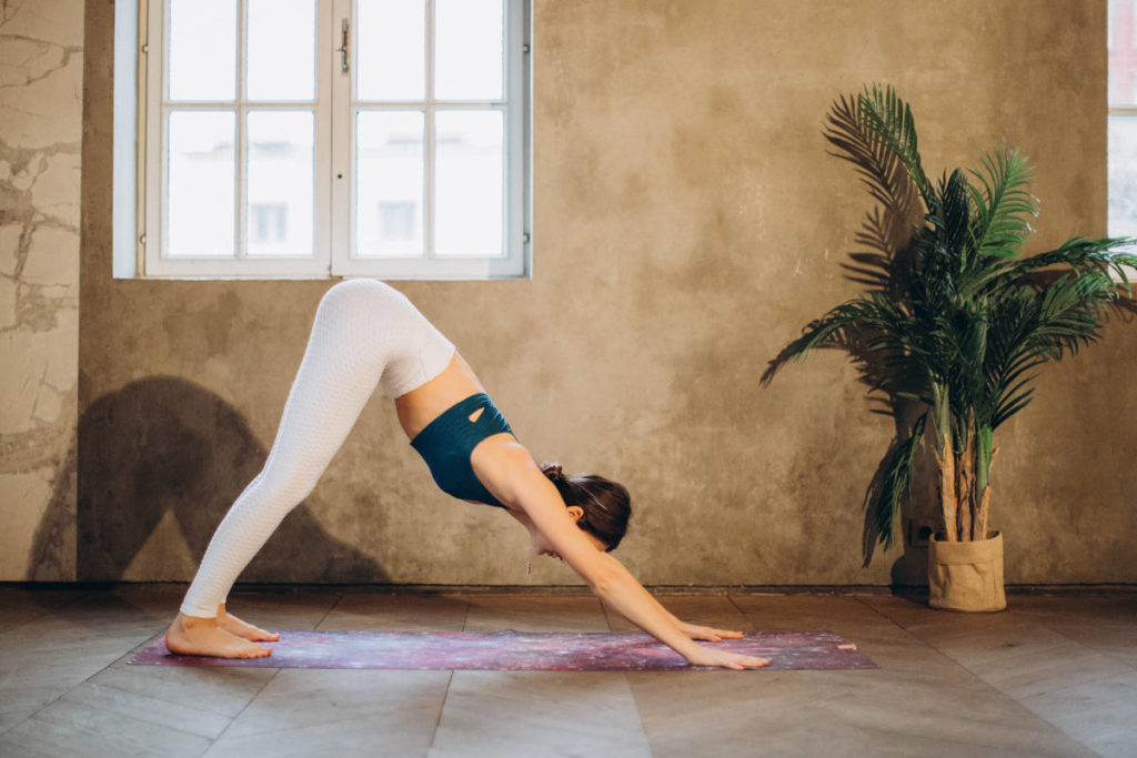 Woman in white leggings and blue top on galaxy mat doing downward facing dog yoga poses for digestion.